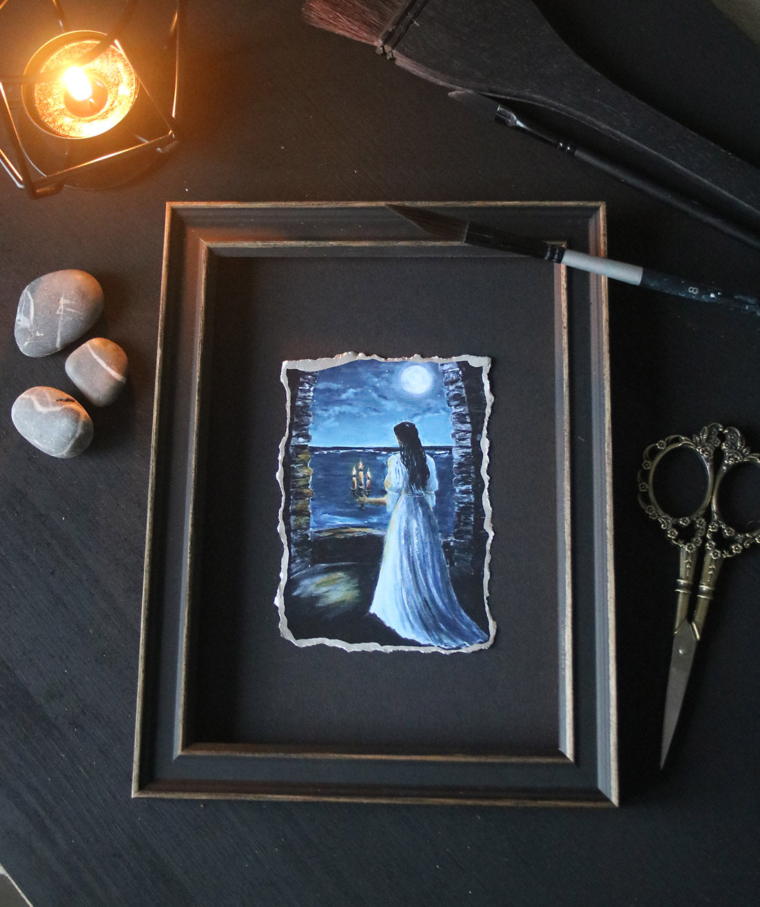 Framed artwork of a woman in a white dress against a dark background, surrounded by decorative items on a dark surface.