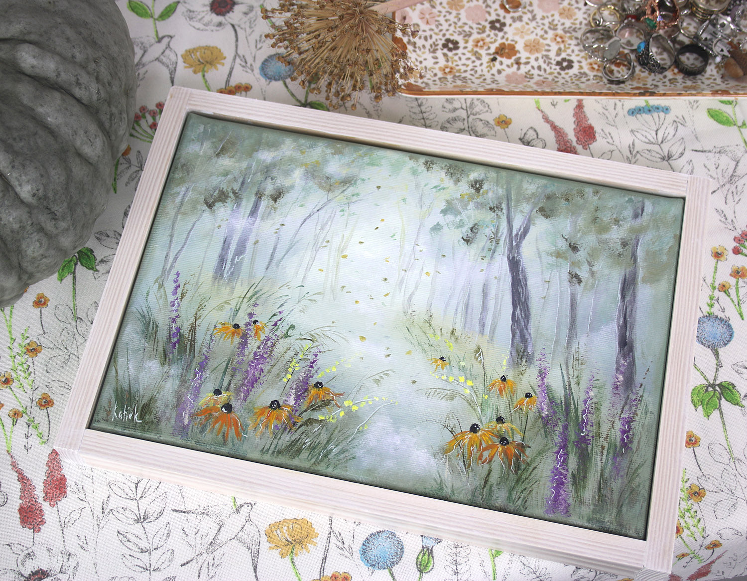 Framed artwork of a nature scene with trees, flowers, on a floral-patterned background.