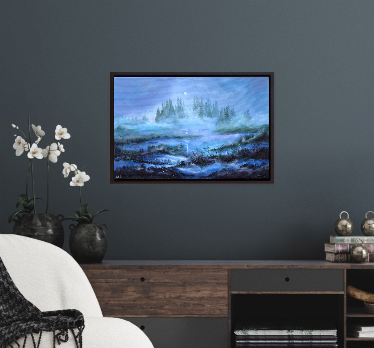 Framed artwork of a landscape with trees and water on a dark gray wall above a wooden shelf.