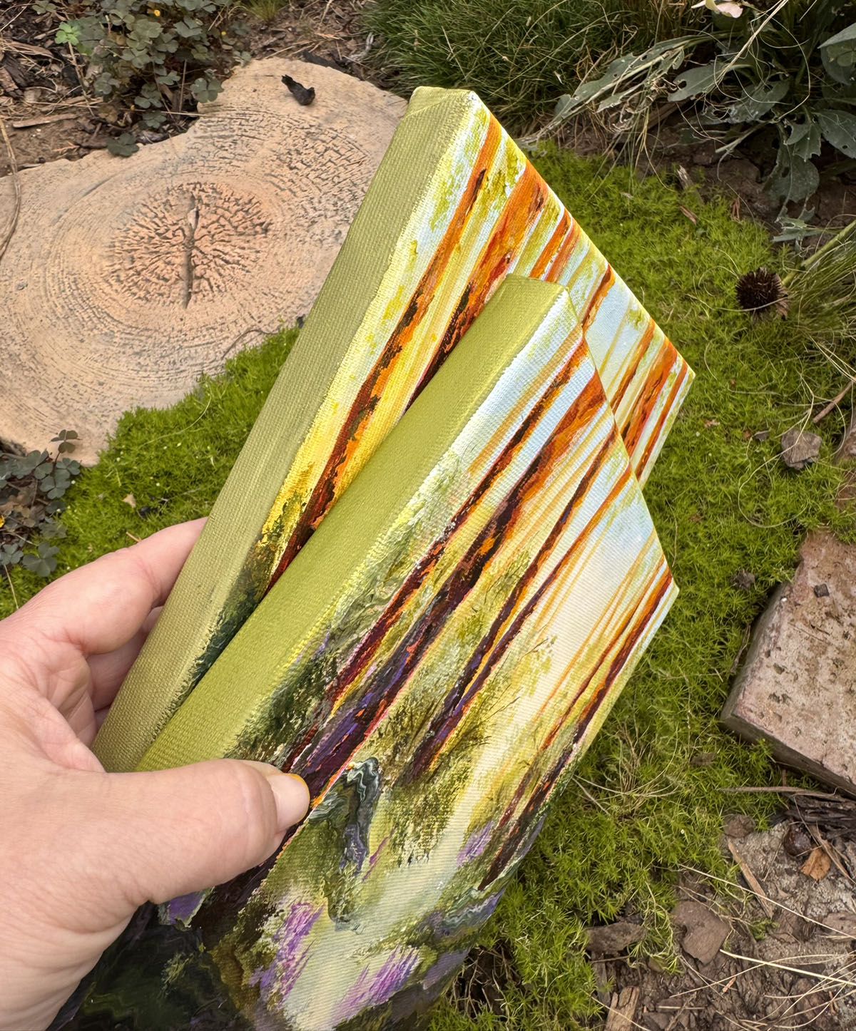 A hand holds two small vertical canvas paintings outdoors over green moss and tree stumps. The canvases depict an abstract landscape with vertical streaks of yellow, orange, brown, purple, and pale blue on an olive-green ground.