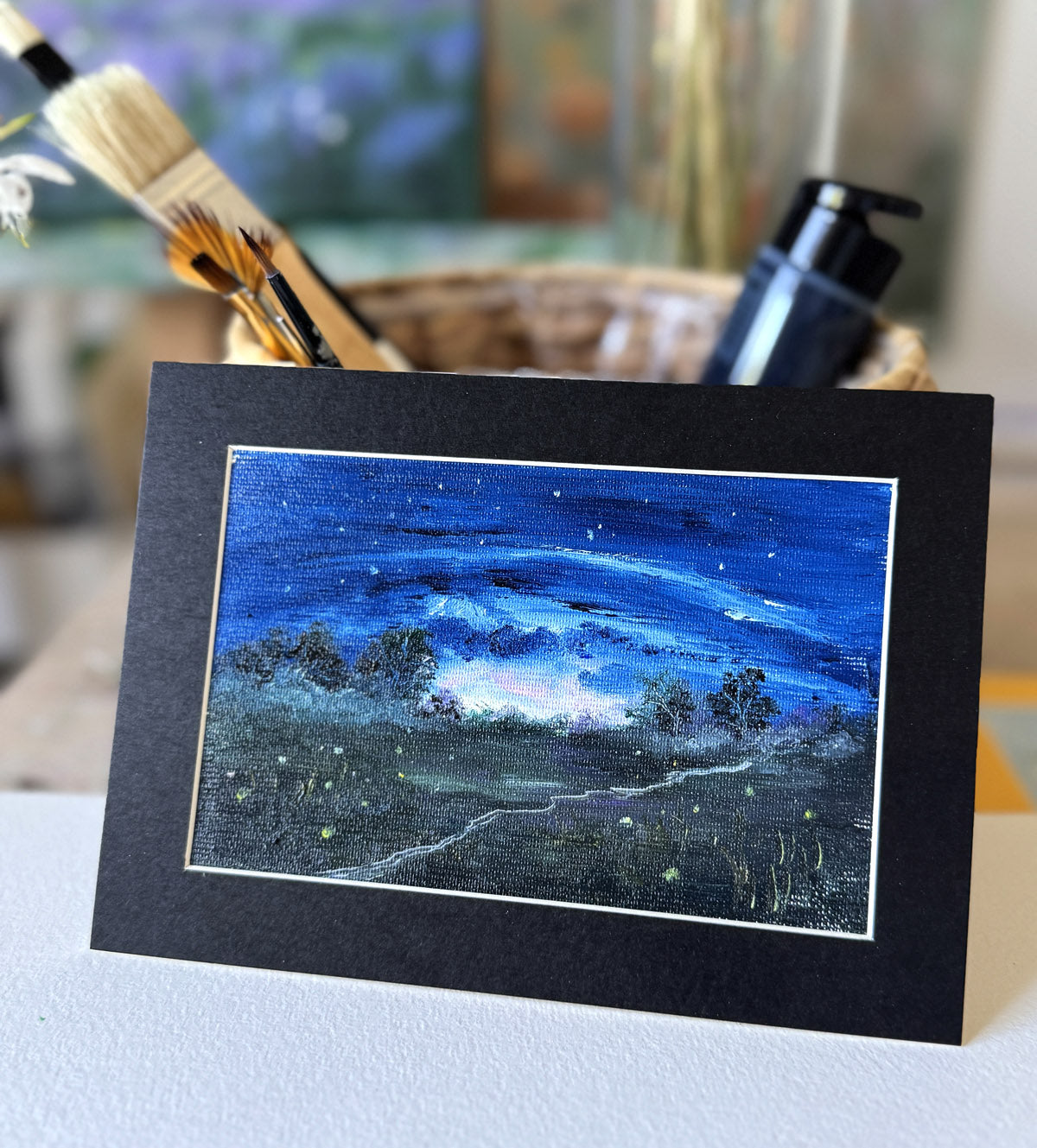 Matted artwork of a night sky scene with trees and stars, placed on a light blue surface.