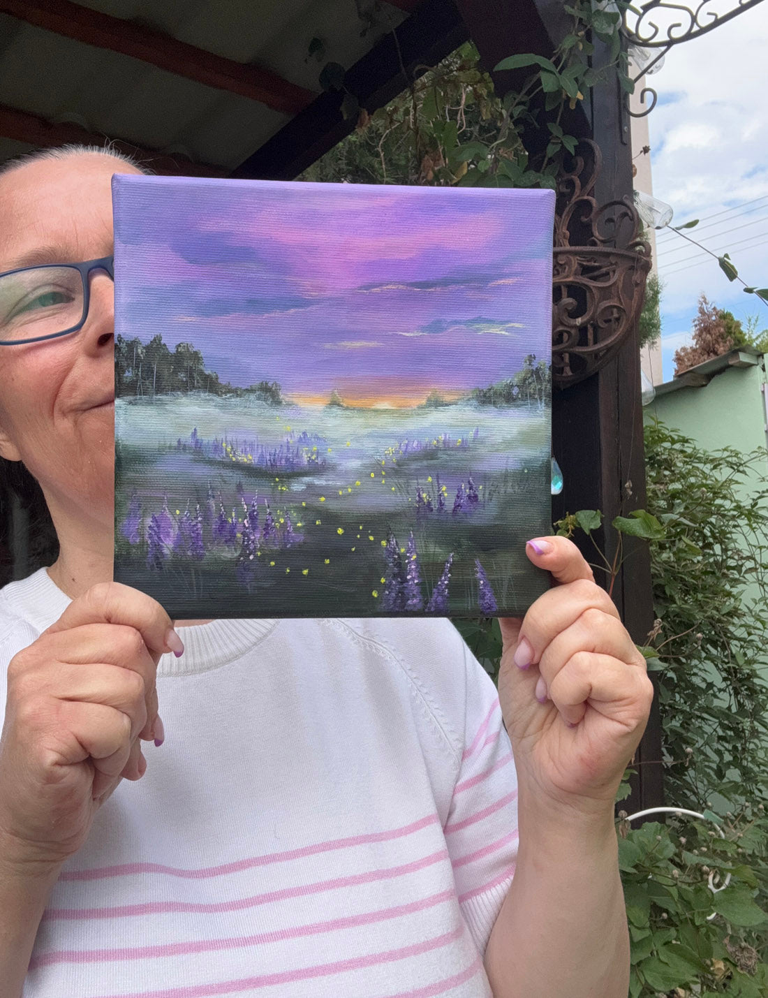 A painting of a misty meadow at dusk with fireflies, with purple and lavender flowers in the foreground, dark tree silhouettes on both sides, and a pink, purple, and blue sky above.