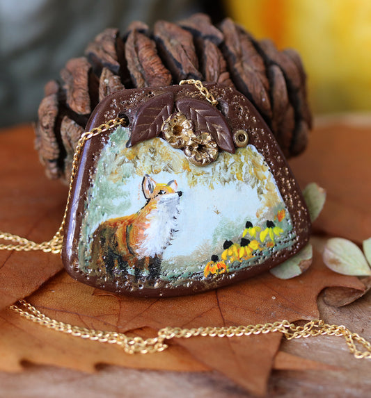 Necklace with a pendant featuring a painted fox on a rock, surrounded by pine cones and leaves.