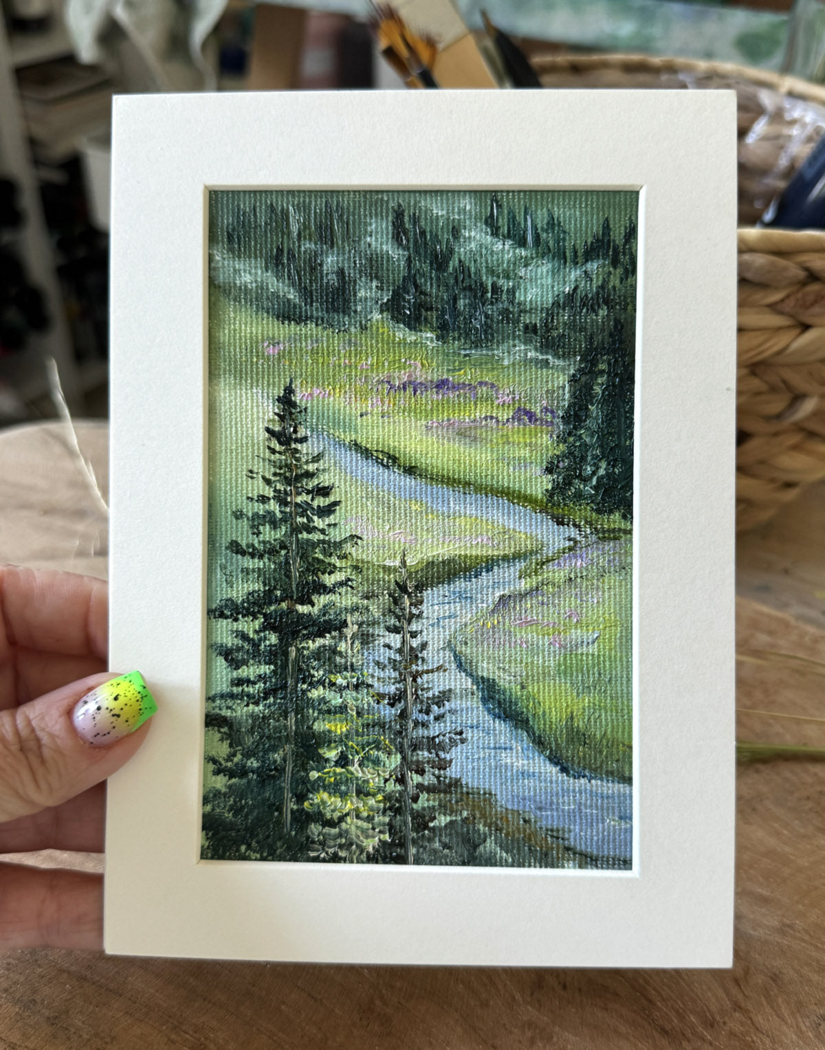 Hand holding a small painting of a landscape with trees and a river.