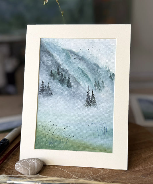 Framed artwork of a mountain landscape with trees on a wooden surface.