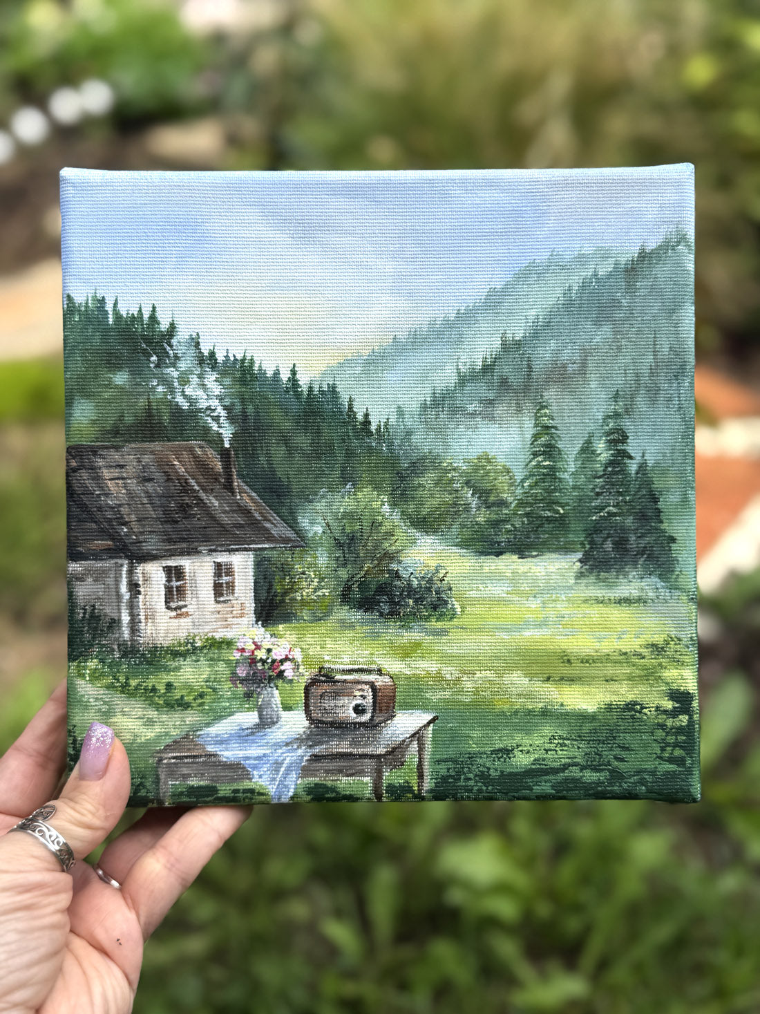 Hand holding a small painting of a cabin in the woods with a radio and flowers on a table.