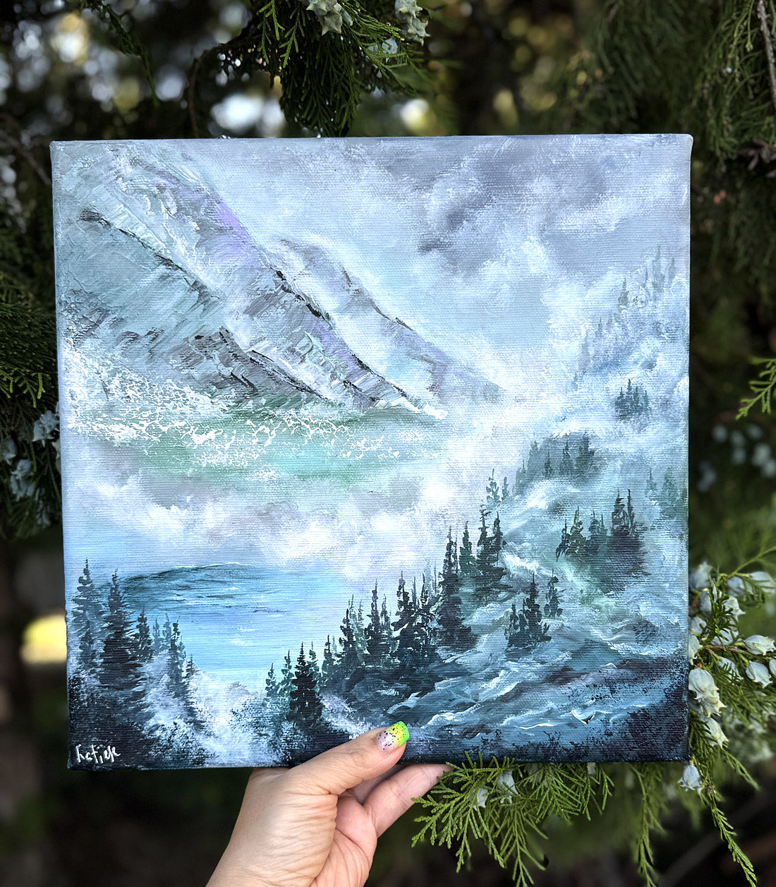 Hand holding a painting of a snowy mountain landscape with trees.