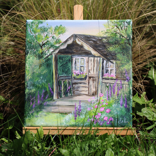 Painting of a wooden cabin in a garden with flowers on an easel surrounded by grass.