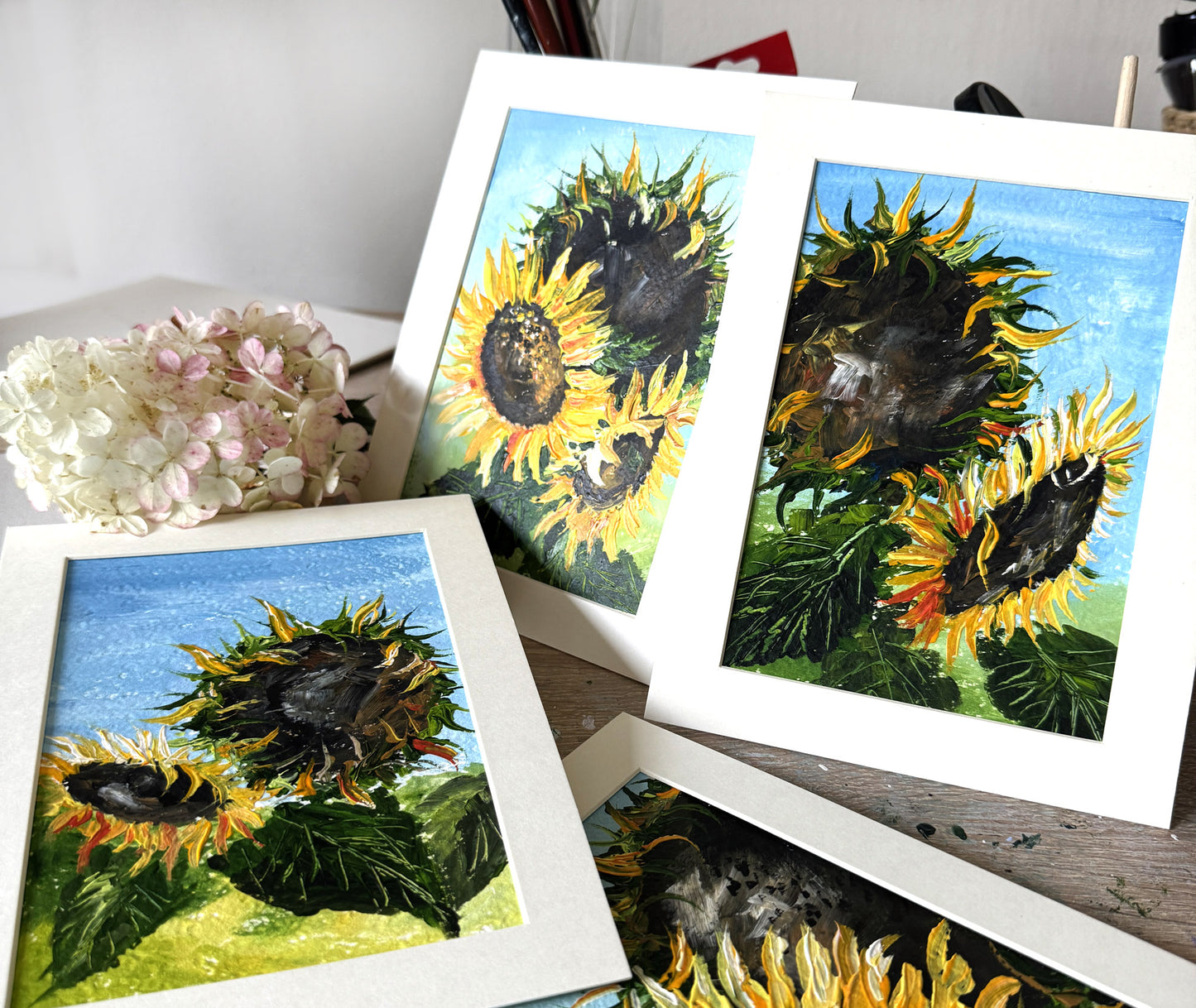 Several matted paintings featuring painted sunflower scenes are arranged on a tabletop, with a bouquet of pale pink hydrangeas beside them.