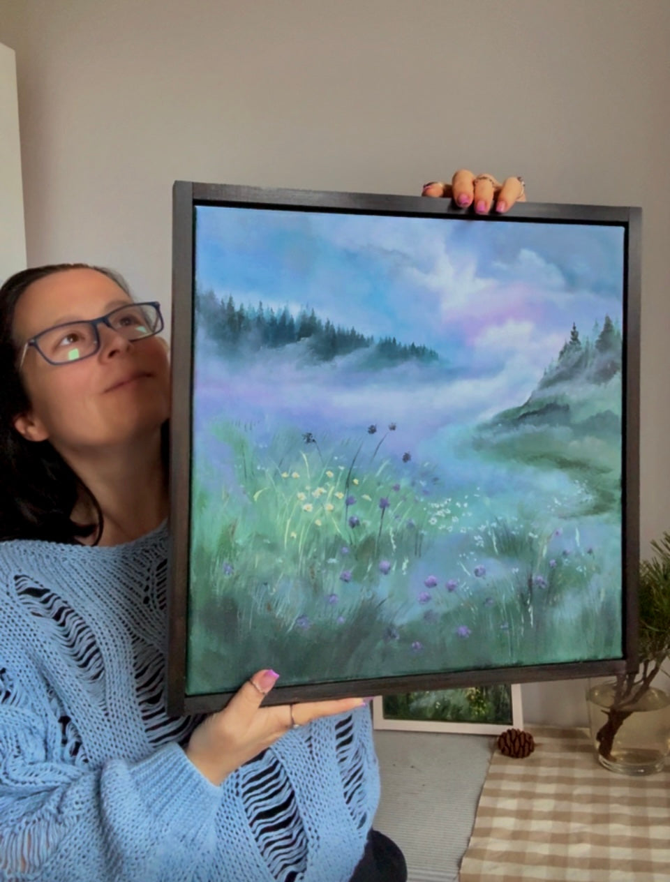 Person holding a framed painting of a landscape with trees and a path.