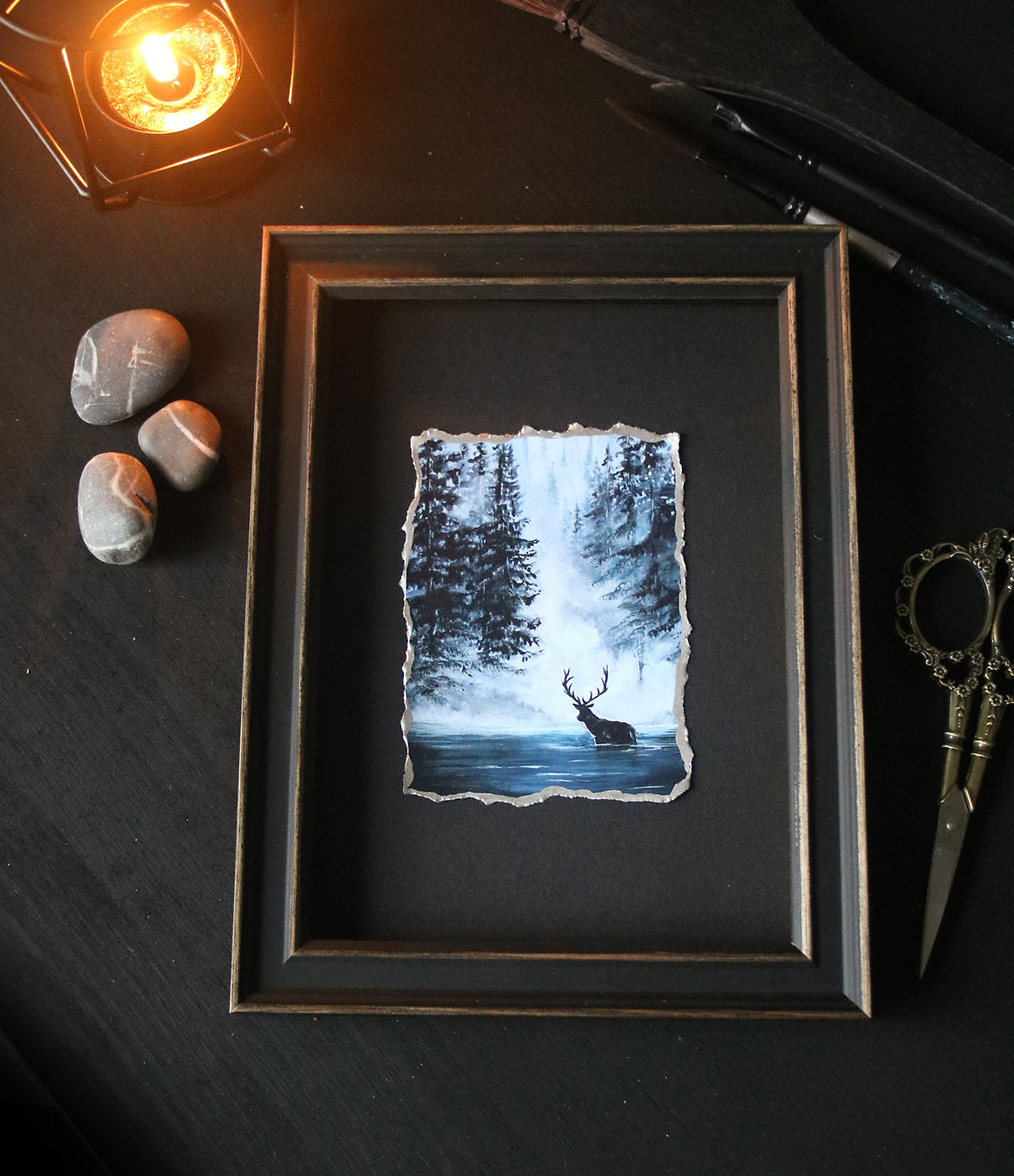 Framed artwork of a stag in a forest with stones and scissors on a dark surface