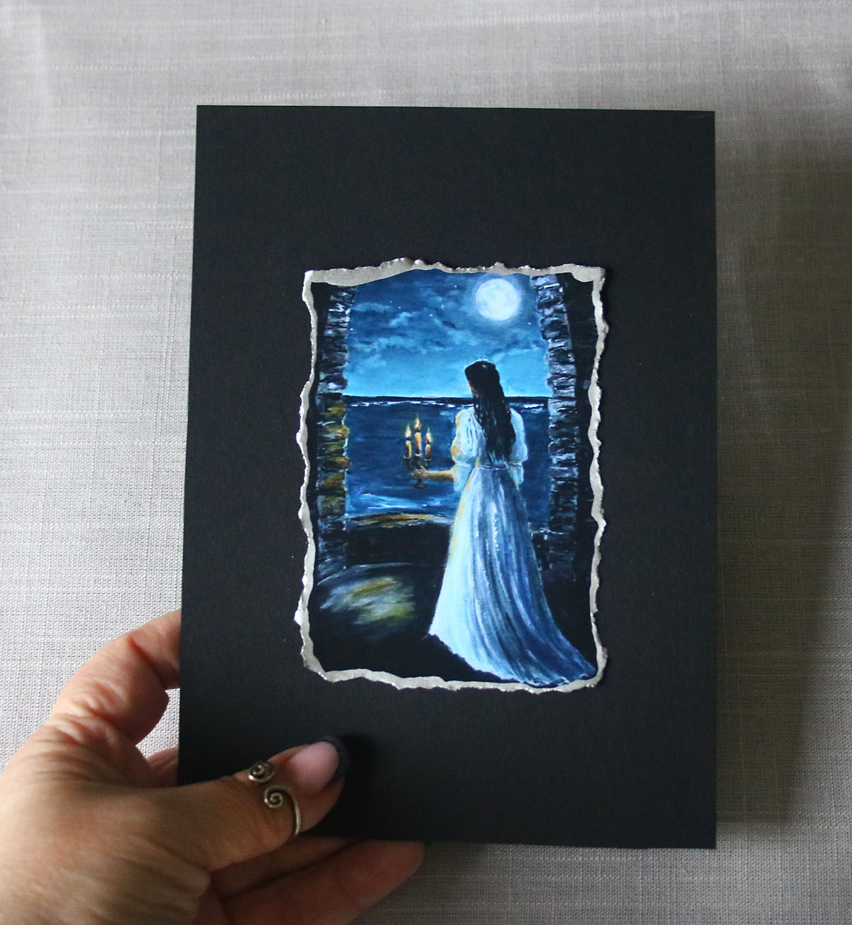 Hand holding a black card with a painted scene of a woman in a blue dress by a window at night.