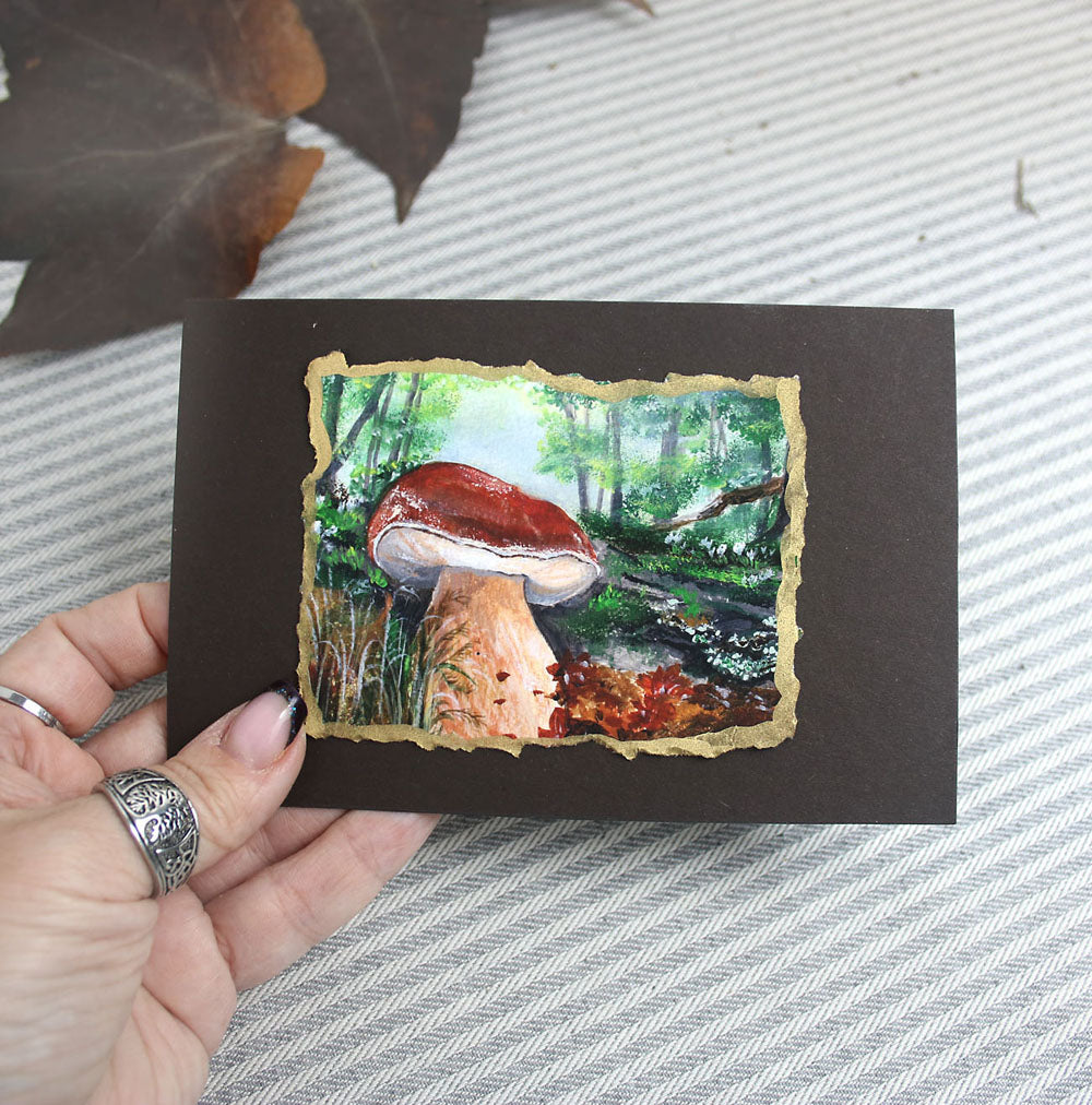 Hand holding a small painting of a forest scene with a mushroom on a textured surface.