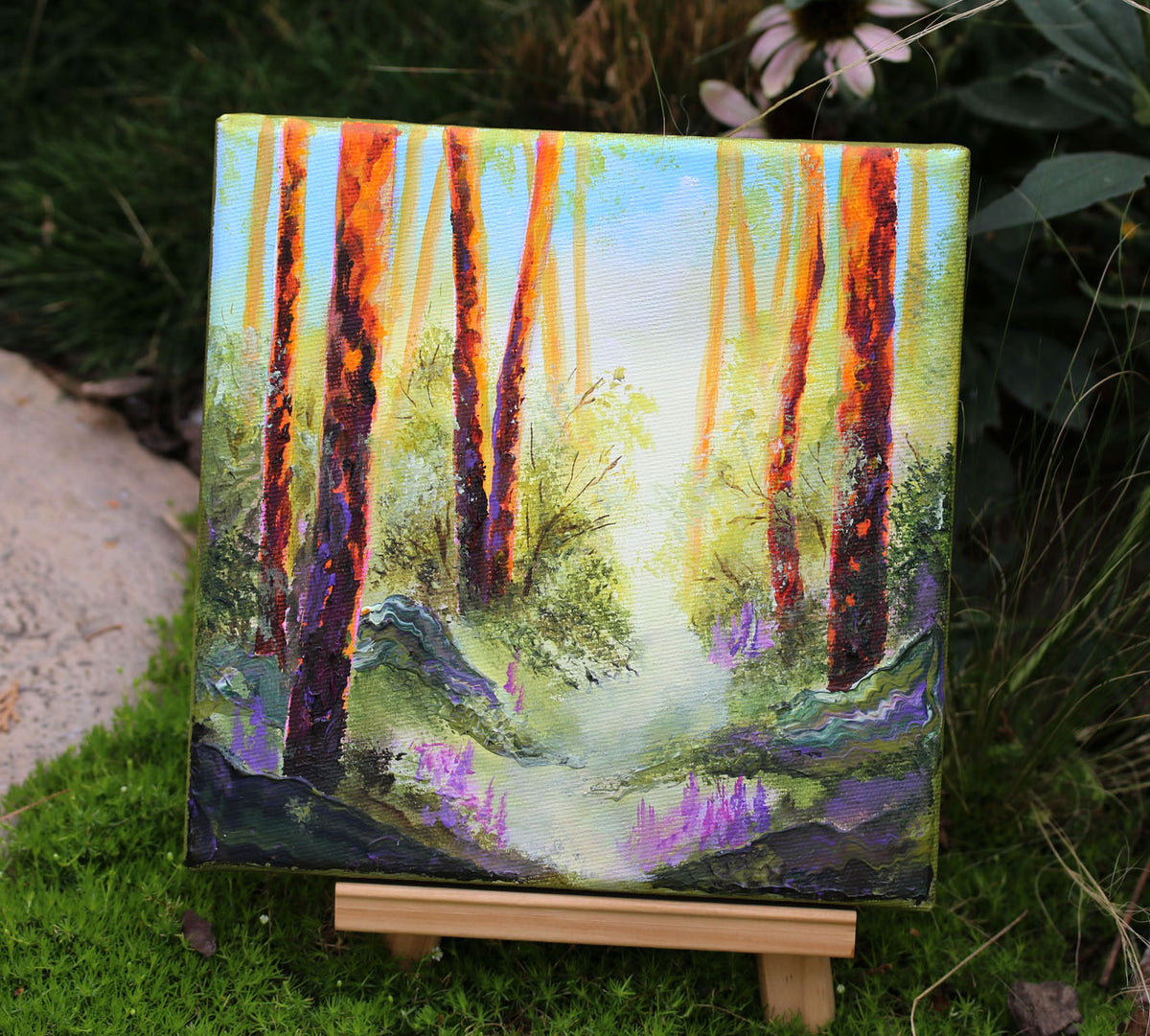 A square canvas painting of a forest path with tall orange and red tree trunks, green foliage, and purple ground reflections, displayed on a small wooden easel outdoors on grass with plants in the background.