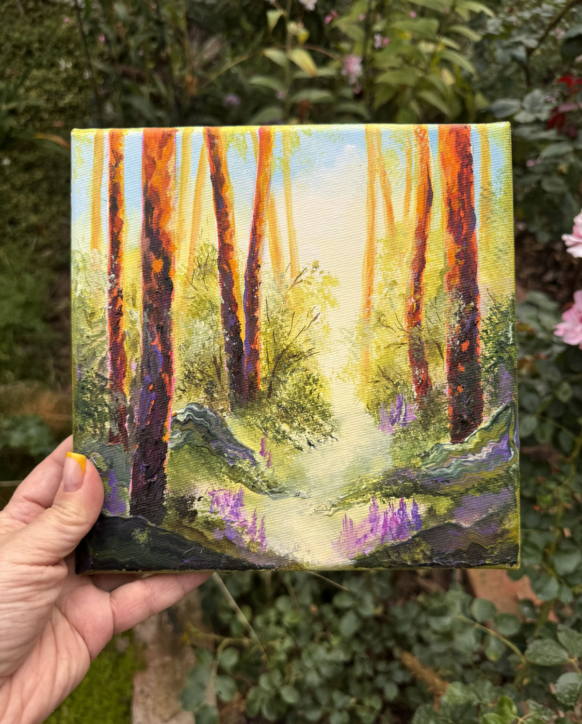 A hand holds a small square canvas painting of a sunlit forest path with tall orange and brown tree trunks, green foliage, and purple flowers, against a real garden background.