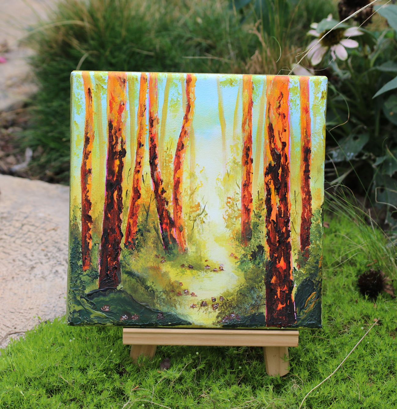 A square canvas painting of a sunlit forest path with tall orange and red tree trunks, displayed on a small wooden easel outdoors on grass with plants in the background.