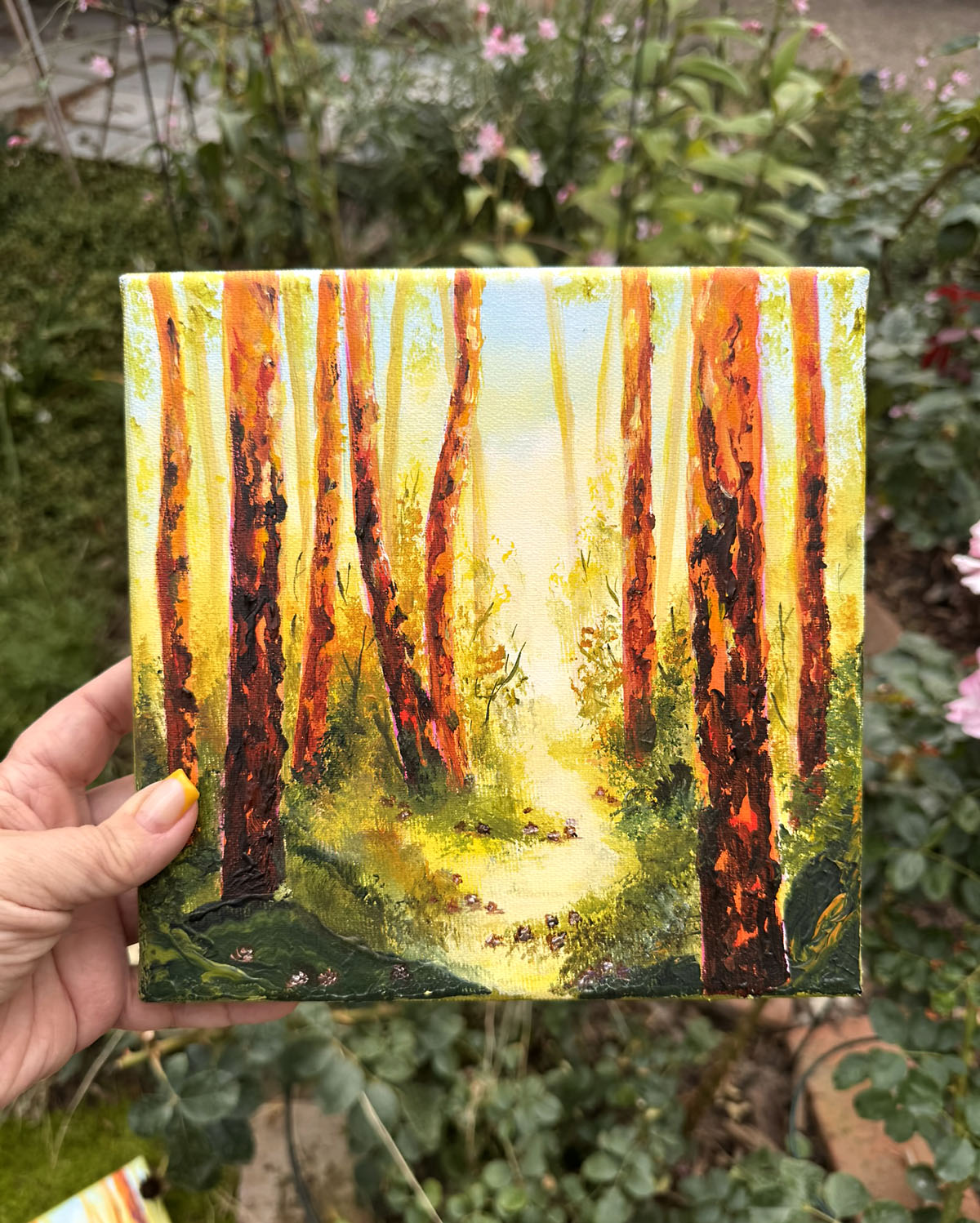 A hand holds a small square canvas painting of a sunlit forest path with tall orange and brown tree trunks, photographed outdoors in front of green garden foliage.