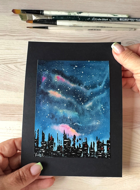 Hand holding a small painting of a cityscape under a starry night sky.