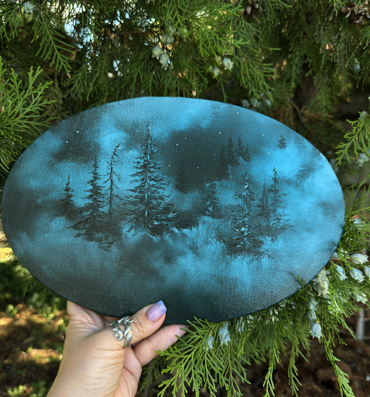 Oval-shaped blue painting with forest design held in front of green foliage