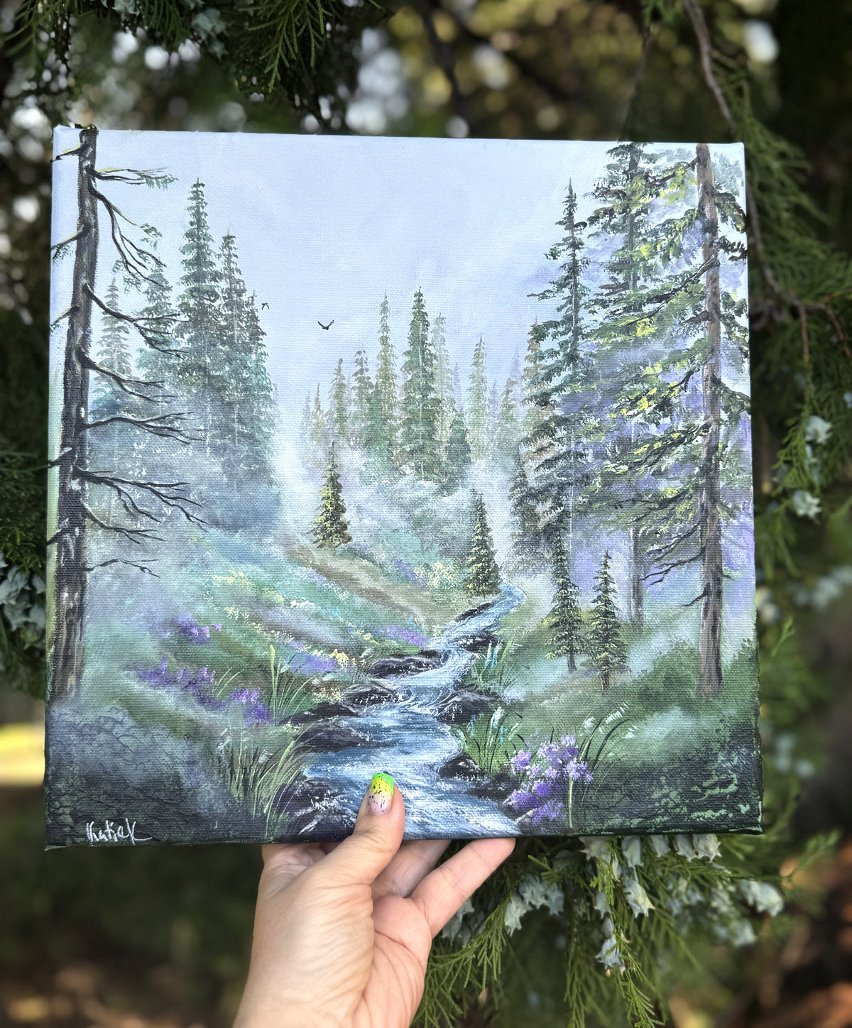 Hand holding a painting of a forest scene with a stream and purple flowers.