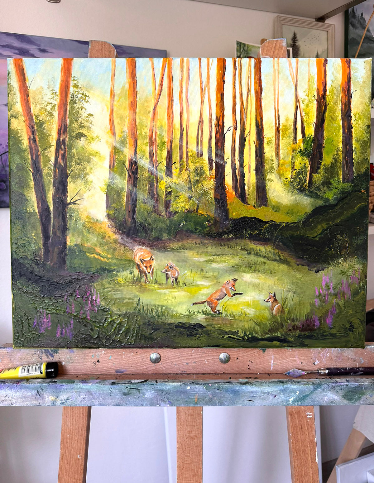 A square canvas painting of a sunlit forest clearing with foxes, displayed on an easel. The artwork shows tall trees, green grass, purple flowers, and warm golden light, with a paint tube resting below the canvas.