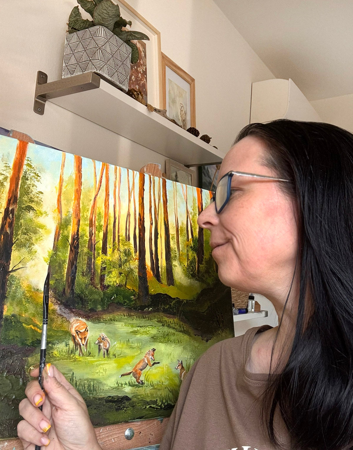 A woman with long dark hair and glasses holds a paintbrush while looking at a large canvas painting of a forest scene with deer. The painting is displayed on an easel in a home studio setting with shelves and framed art in the background.