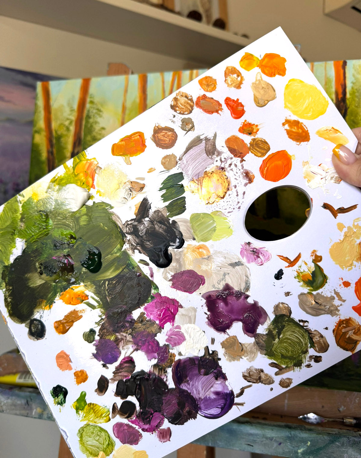A white artist's paint palette covered with thick, textured paint swatches in various colors, held up in an art studio setting.