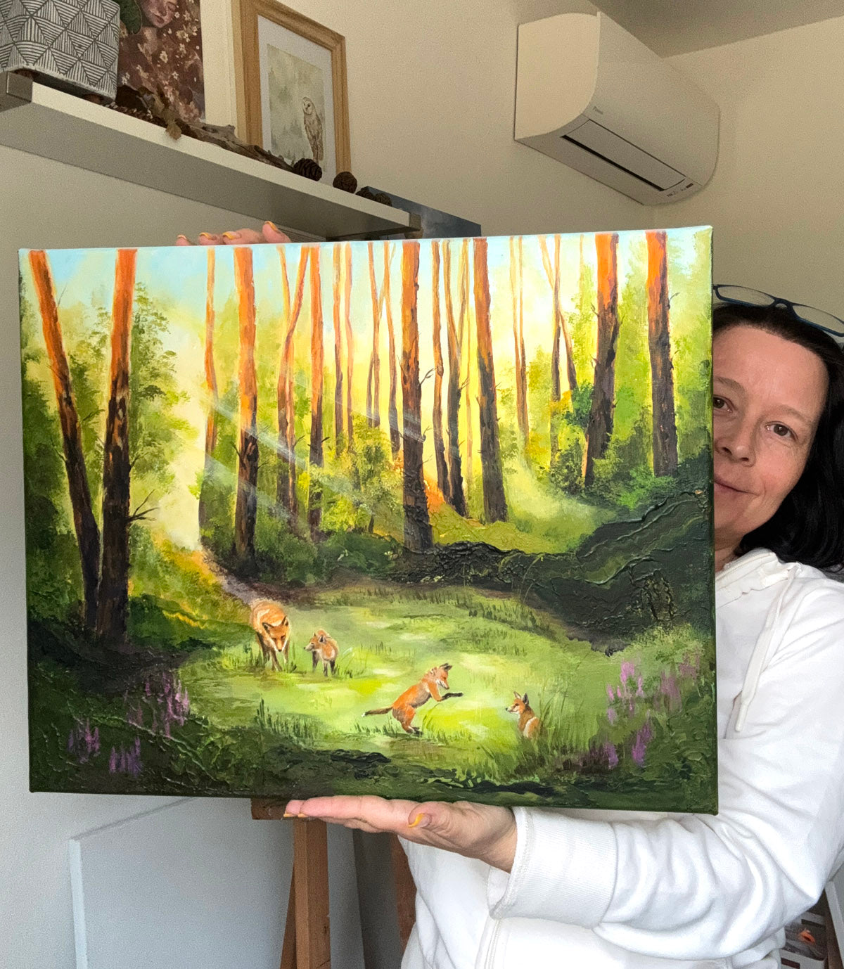 A woman in a white hoodie holds up a colorful oil painting of a forest scene with foxes on the ground, set against a home interior background with a visible air conditioner and shelf.