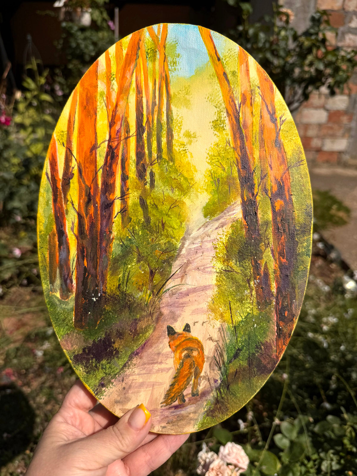 An oval hand-painted wooden plaque depicting a fox walking along a path in a sunlit forest, held by a hand against a blurred garden background.