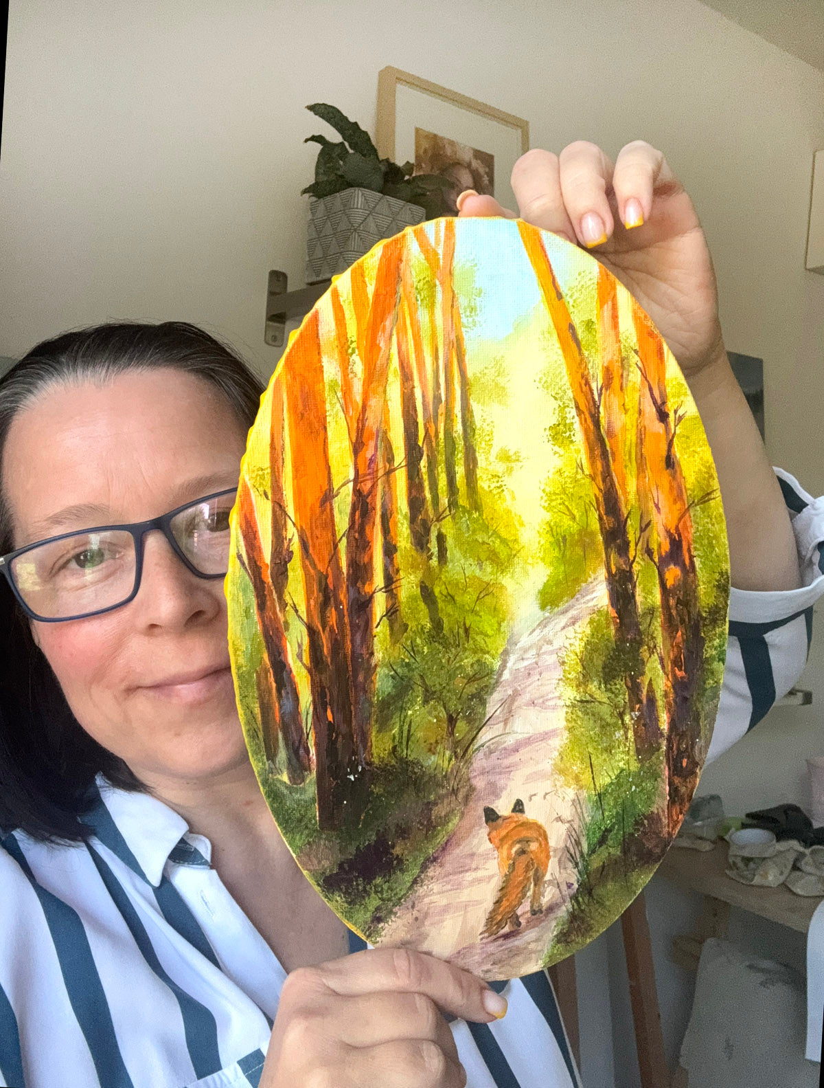 A woman holding up an oval-shaped acrylic painting of a forest path with orange trees and a fox, in an indoor setting.