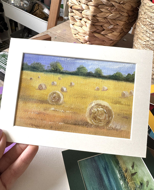 A hand holds a framed landscape art print depicting a golden field with hay bales under a blue sky. The print features a textured, woven canvas look and is presented in a white mat board frame.