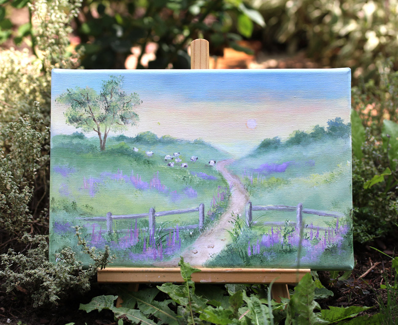 Painting of a landscape with a path, sheep, trees, and sheep on an easel surrounded by greenery.
