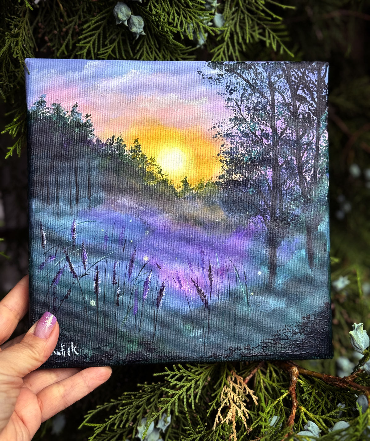 Hand holding a small canvas with a dreamy sunset landscape painting in purple and green tones.