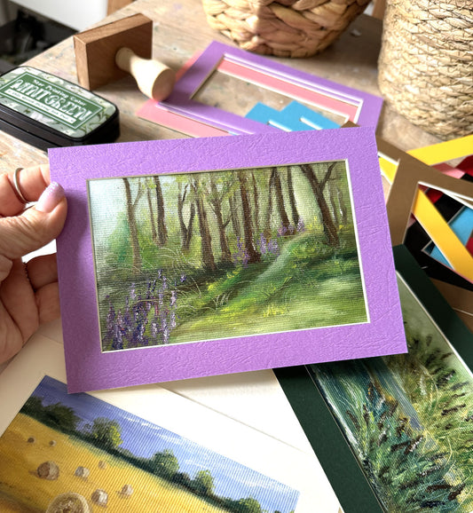 A hand holds a square art print featuring a painted woodland landscape with a purple border, surrounded by craft supplies and other prints on a wooden table.
