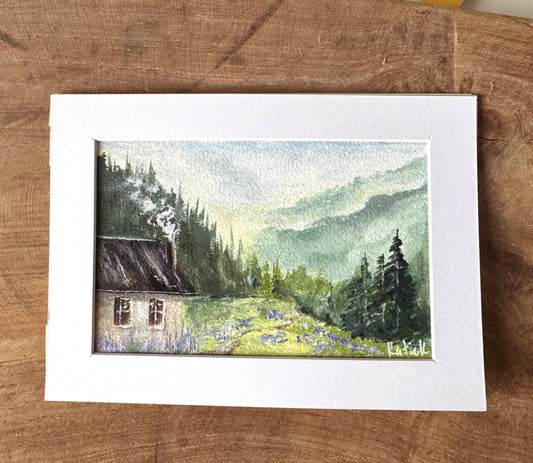 Framed watercolor painting of a cabin in the woods on a wooden surface