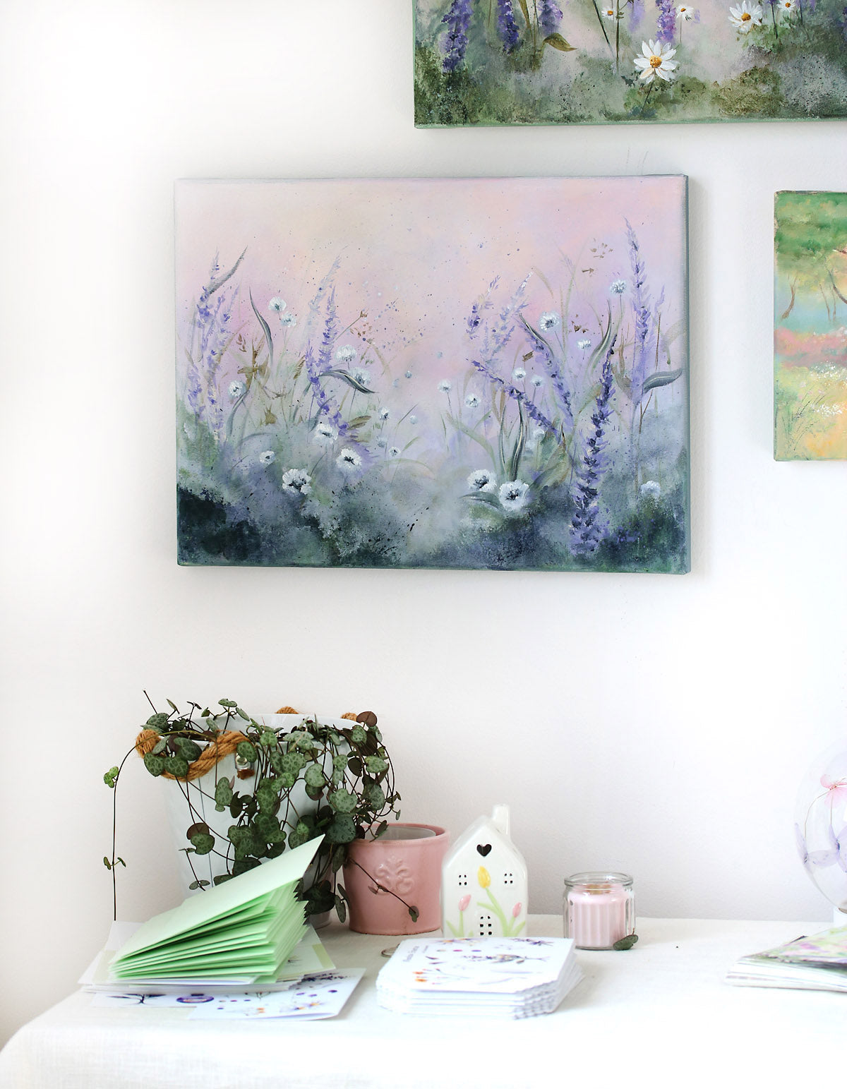 Floral painting on a wall with a table below featuring a plant, candle, and books.