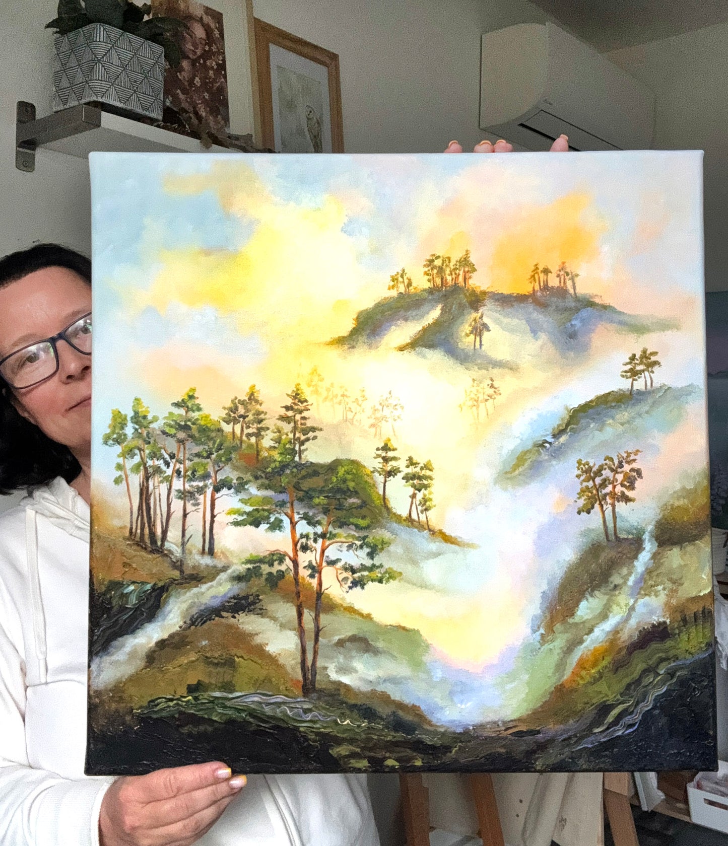 A person holding up a square canvas painting of a misty mountain landscape with trees and a glowing yellow sky. The person is partially visible on the left side of the frame, wearing glasses and a white hoodie, in what appears to be an indoor studio or home environment.