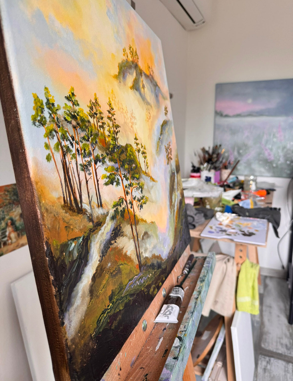 A large vertical canvas painting of a forest landscape with trees, hills, and a waterfall, displayed on an easel in an artist's studio. The painting features warm golden and orange sky tones with green and brown trees.