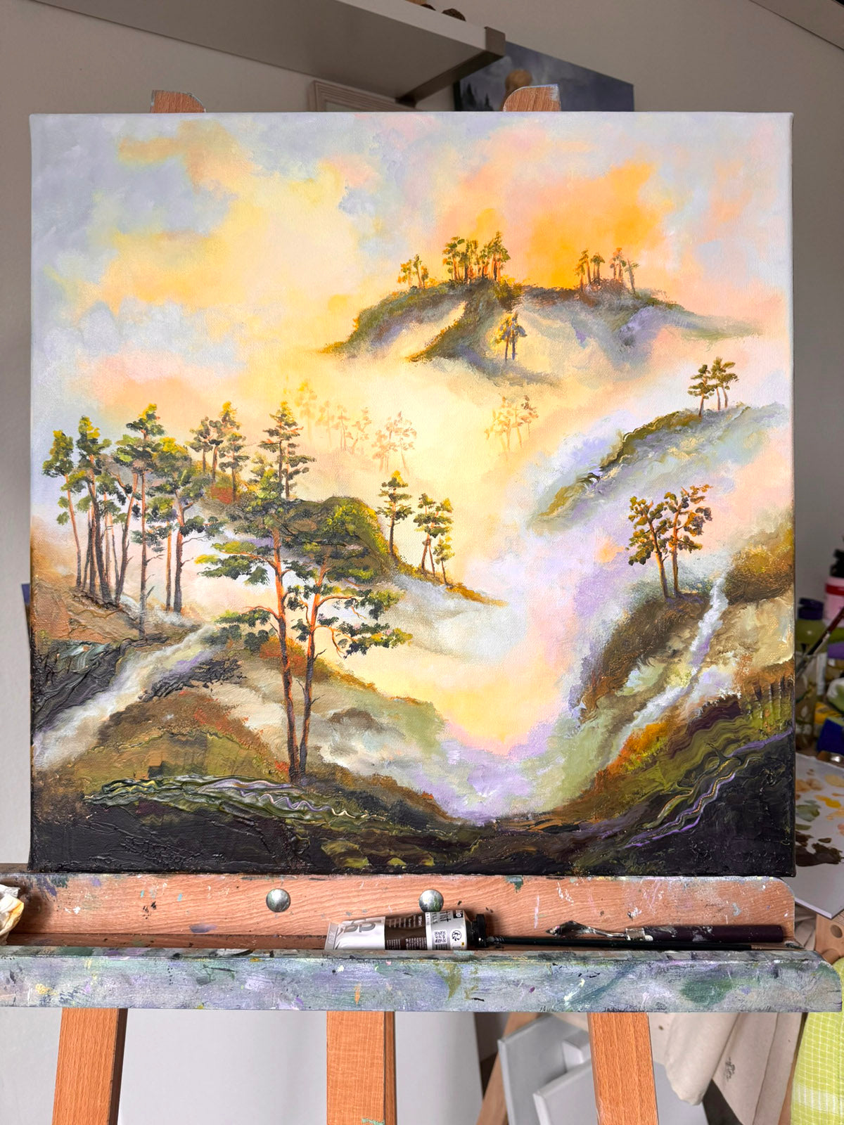 A landscape painting on canvas depicting misty hills, trees, and a glowing yellow-orange sky, displayed on an easel in an artist's studio.