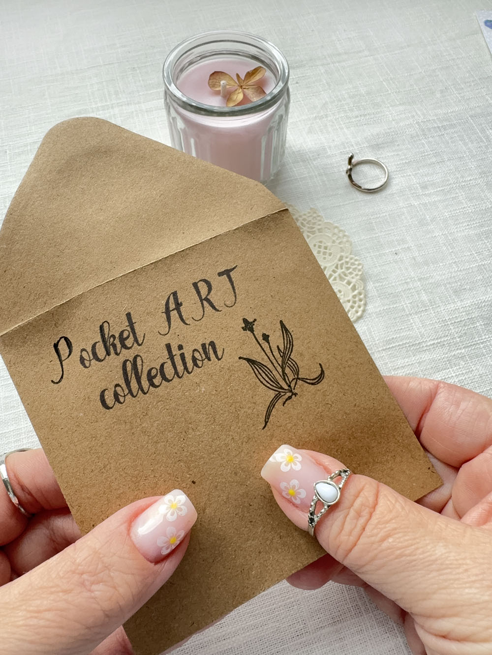 Kraft paper card with 'Pocket Art Collection' text and a small illustration, held by hands with floral nail designs.