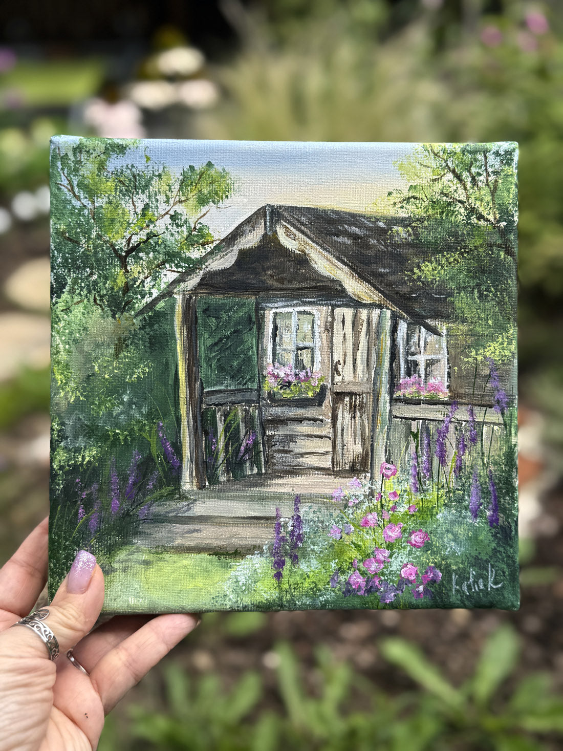 Painting of a wooden shed with flowers in a garden, held by a hand.