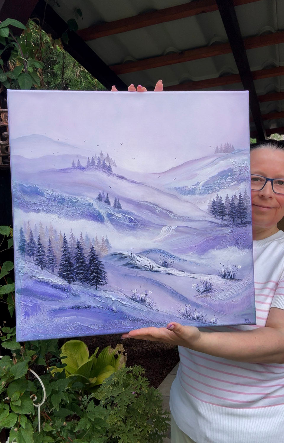 A person holding up a square canvas painting of a misty purple and blue mountain landscape with pine trees, photographed outdoors under a sheltered roof with plants visible around it.