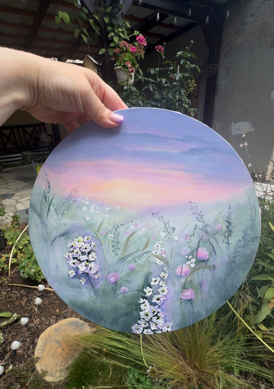 A circular canvas painting featuring a painted meadow scene with white and pink wildflowers under a soft purple and pink sunset sky.
