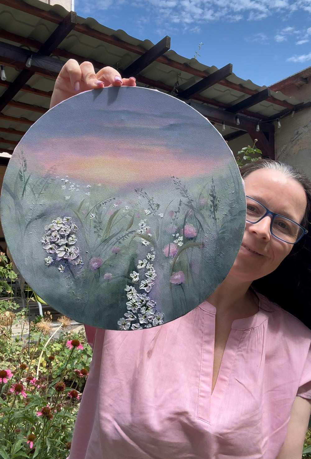 A circular canvas painting featuring a painted meadow scene with white and pink wildflowers under a soft purple and pink sunset sky.