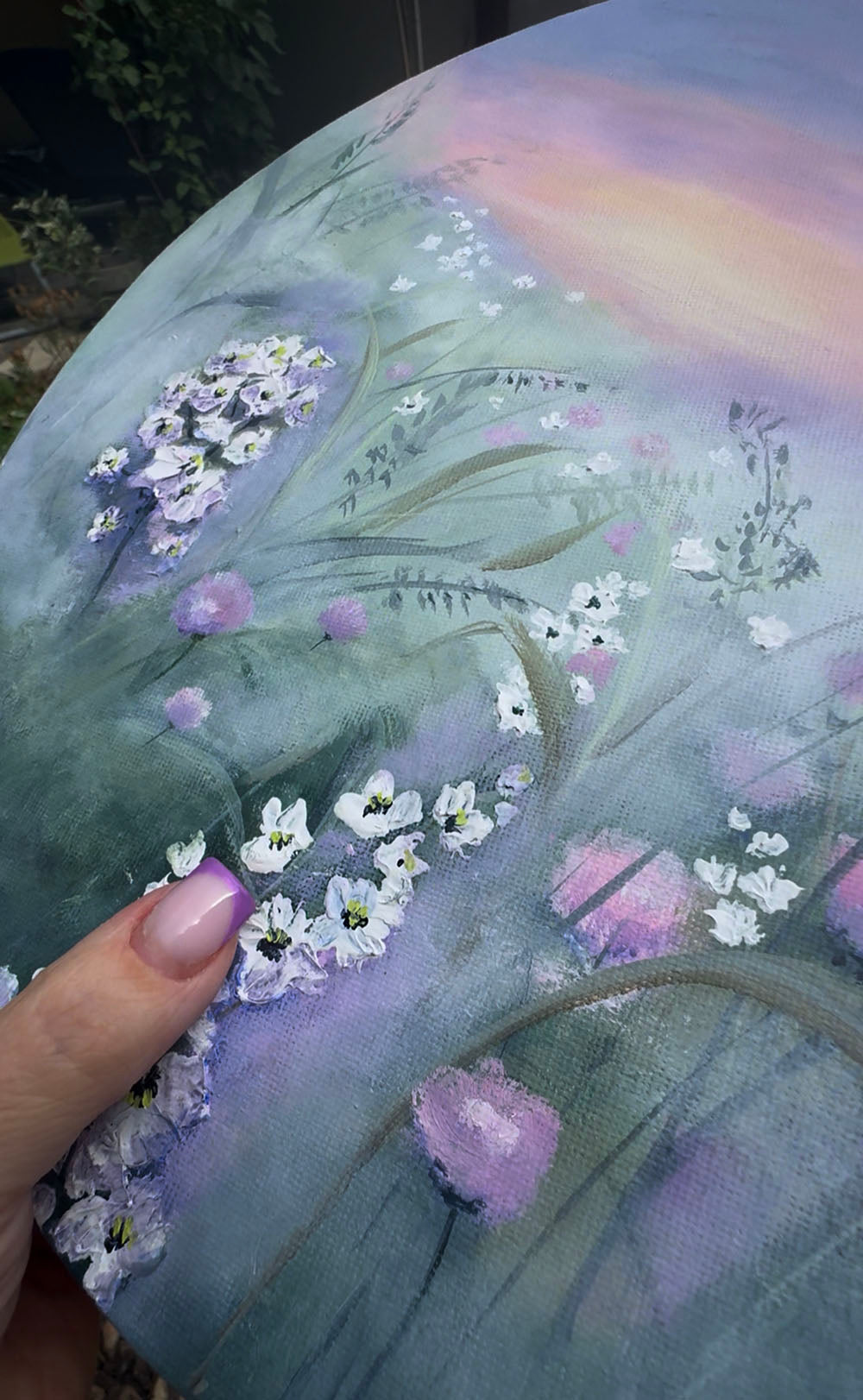 A circular canvas painting featuring a painted meadow scene with white and pink wildflowers under a soft purple and pink sunset sky.