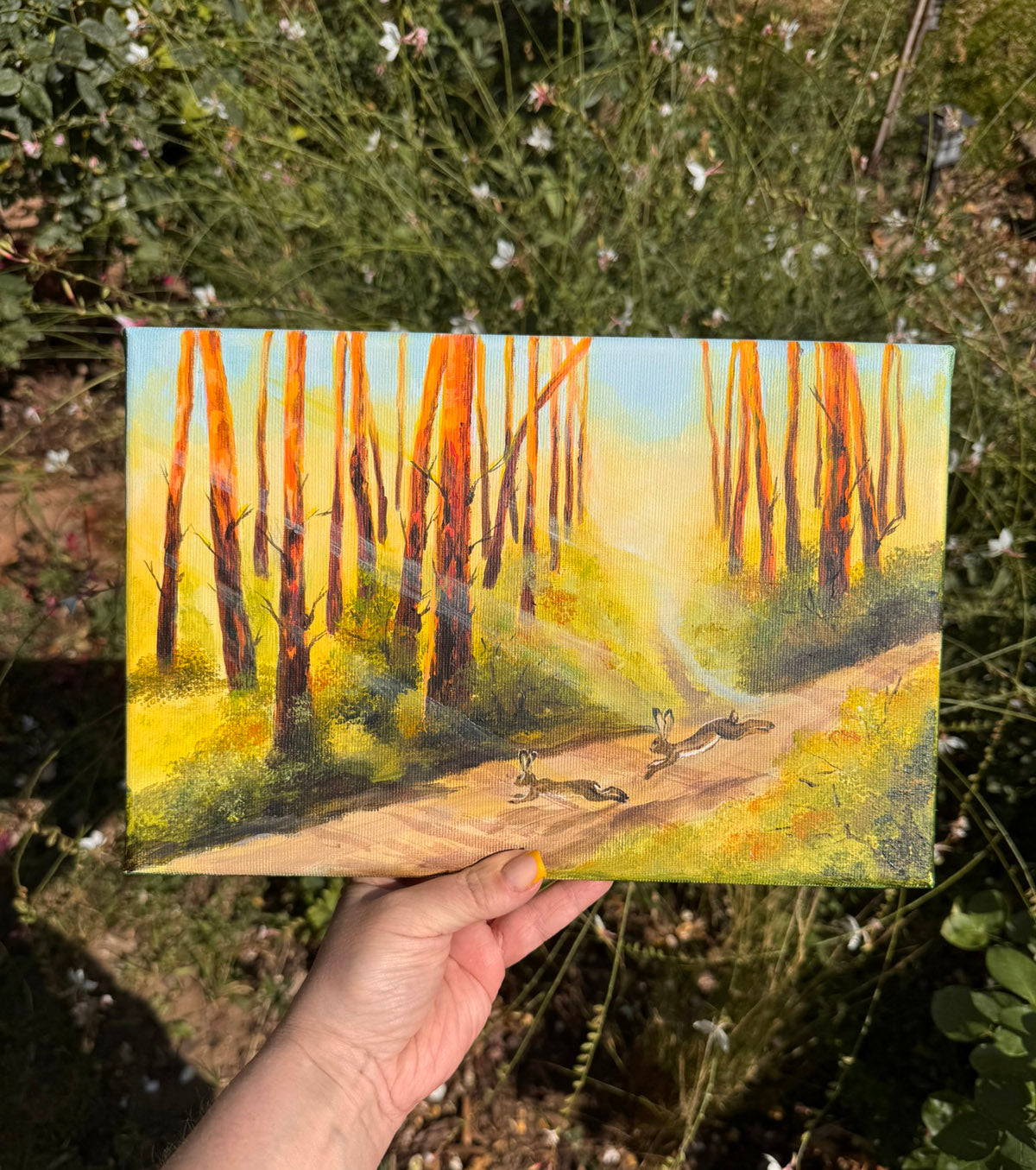 A hand holds a small square painting of a forest path with tall orange-brown tree trunks, yellow-green grass, and two rabbits in a sunlit woodland scene, photographed outdoors against real plants and flowers.