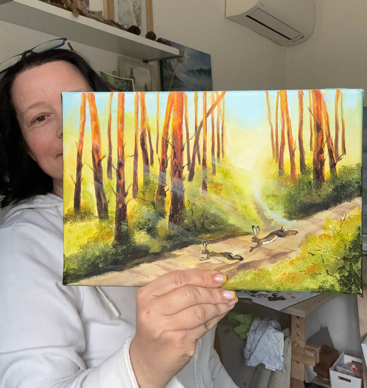 A woman in a white hoodie holds up a square canvas painting of a forest path with tall orange-brown trees and rabbits, shown in a home studio setting.