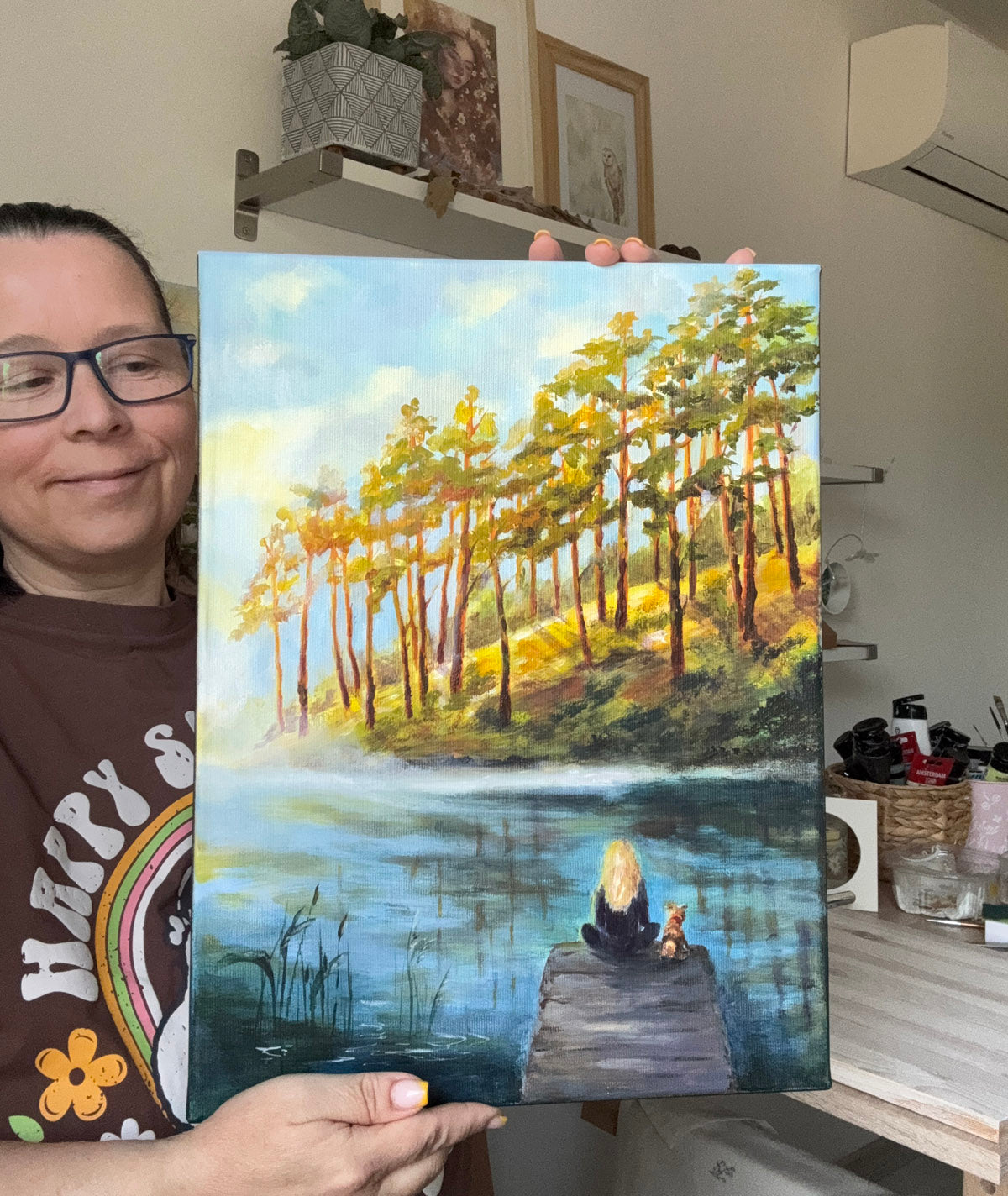 A person holding up a colorful painting of a forest by a lake, with a dock and a small figure in the foreground. The person is partially visible on the left side of the frame, wearing glasses and a brown graphic t-shirt.