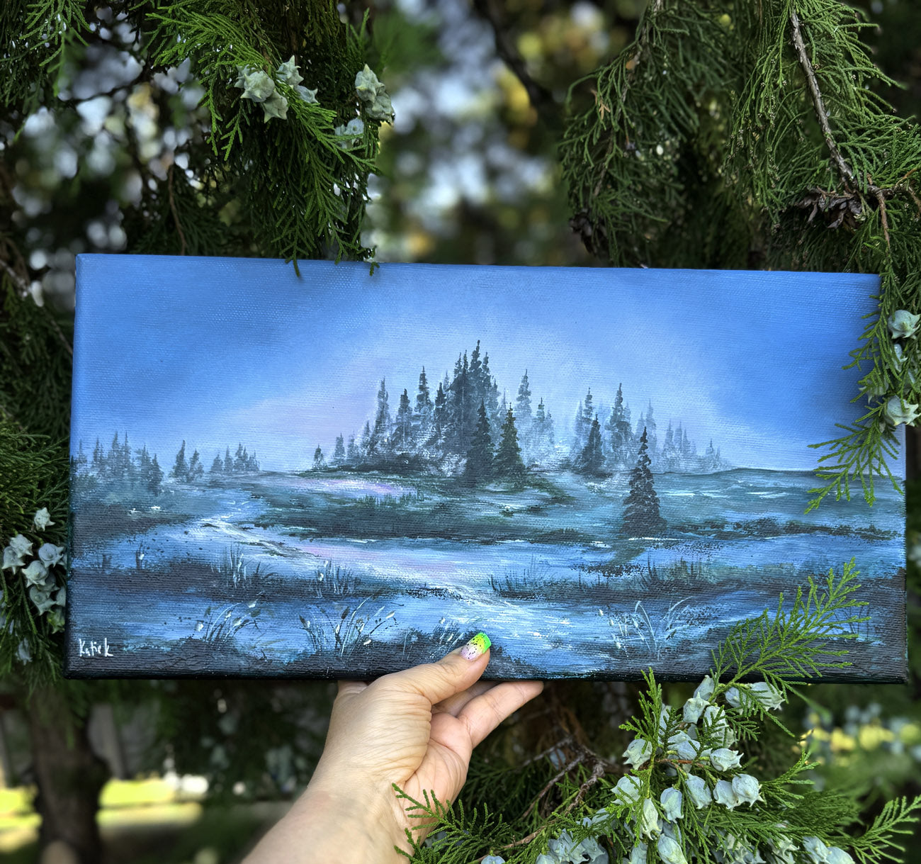 Hand holding a small painting of a forest landscape with a hand holding it against a natural background.