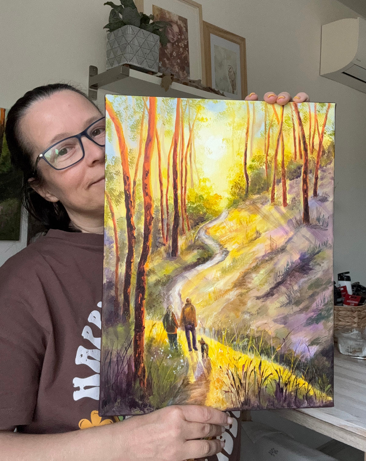 A woman wearing glasses and a brown t-shirt holds up a square canvas painting of a sunlit forest path with tall trees and two small figures walking. The room background includes shelves with framed art and household items.